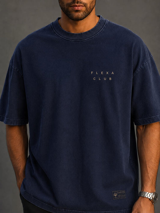 A man wearing a dark navy blue oversized t-shirt with 'FLEXA CLUB' printed in gold on the left chest, photographed against a dark gray studio background.