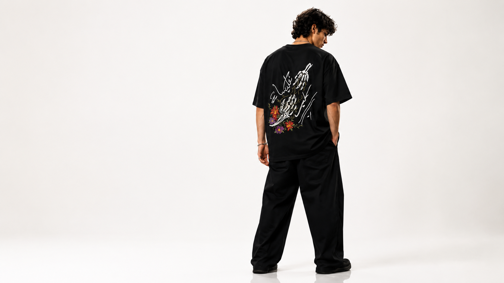 A model viewed from behind wearing a black oversized t-shirt with a large white graphic print and matching black sweatpants, standing against a plain white studio background.
