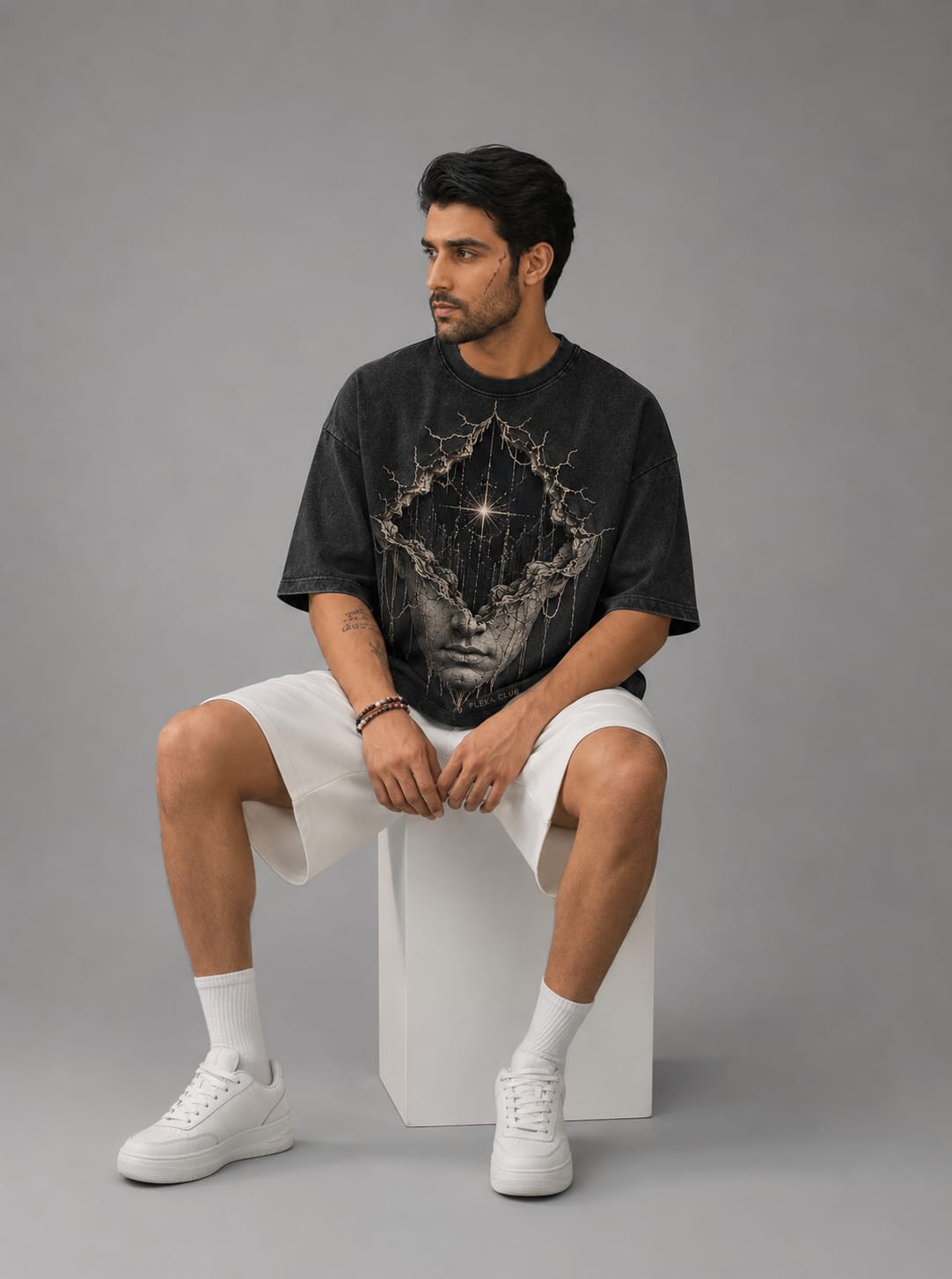 A model shown sitting down on white stool, on a light gray background, wearing a dark gray acid washed drop shoulder T-Shirt with a cosmic themed white sculpture of a face.