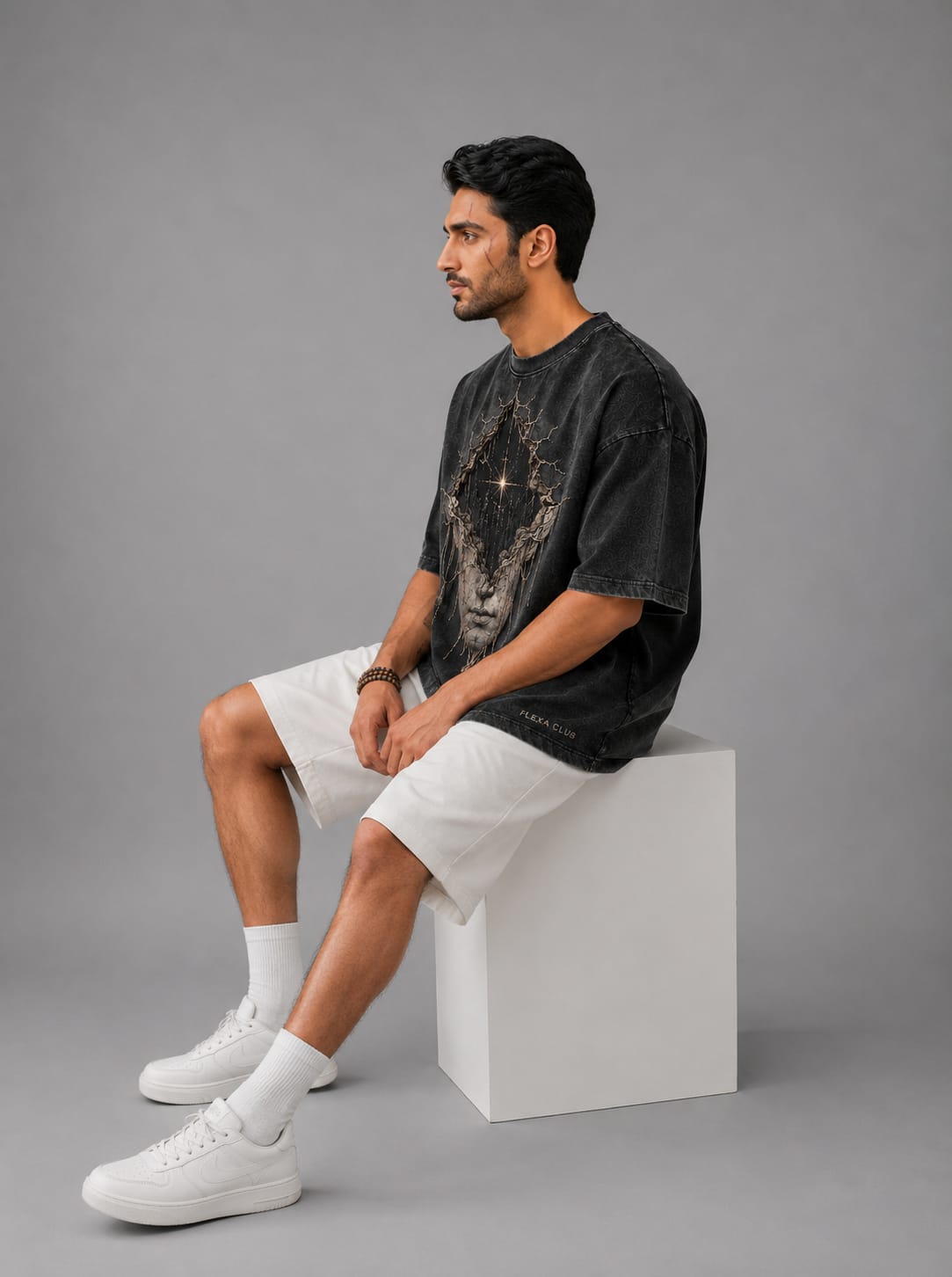 A model shown sitting down from the side on white stool, on a light gray background, wearing a dark gray acid washed drop shoulder T-Shirt with a cosmic themed white sculpture of a face.