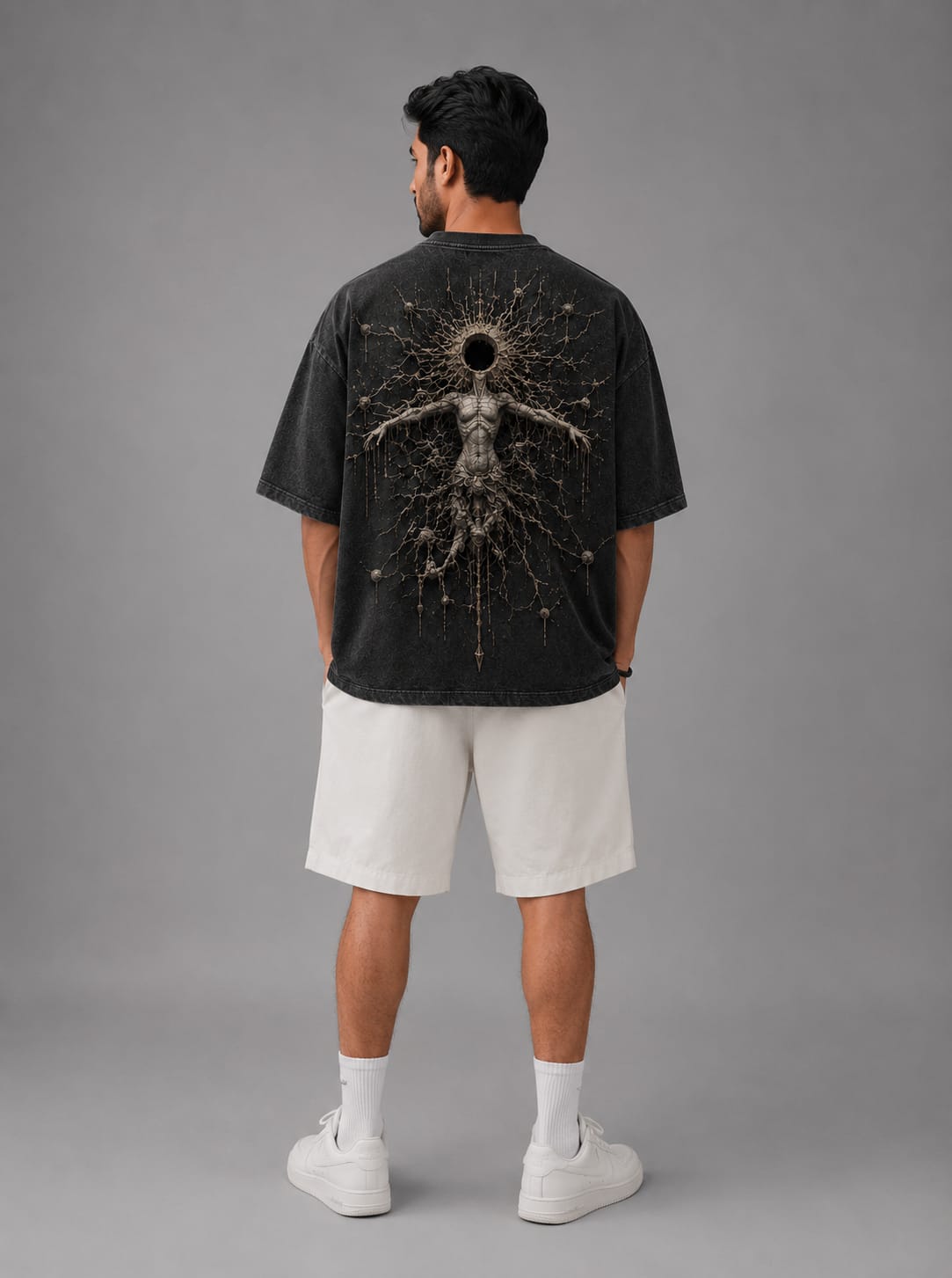 A model shown standing from the back against alight gray background, wearing a dark gray acid washed drop shoulder T-Shirt with a cosmic themed white body sculpture of an entity.