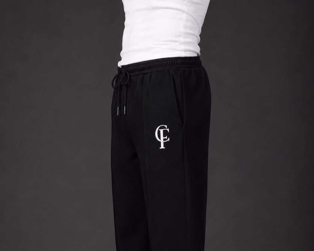 Black Trouser with a white DTF FlexaClub monogram logo on the left thigh, shown on a model wearing a white shirt and white sneakers against a dark gray studio background.