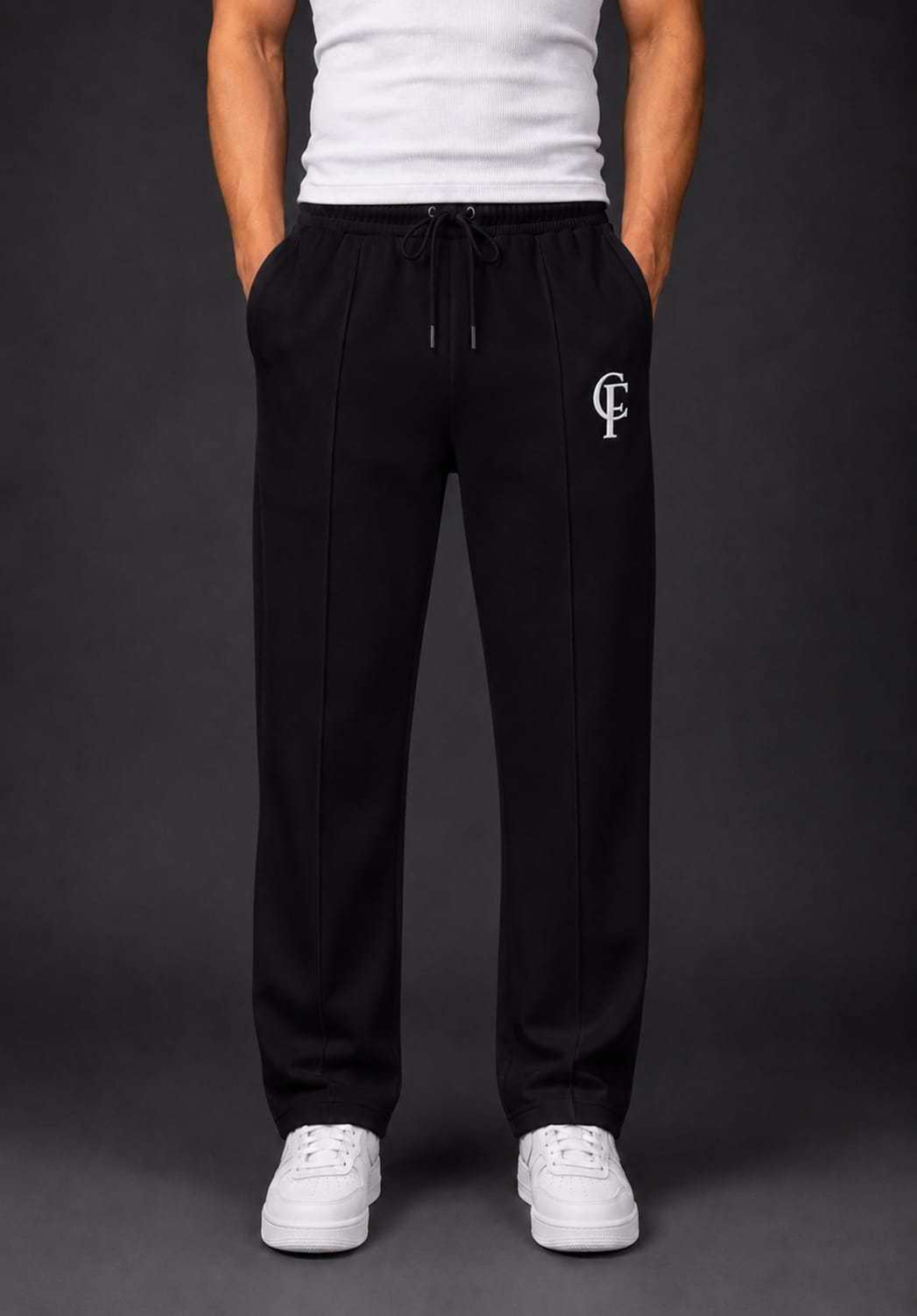Black Trouser with a white DTF FlexaClub "FC" monogram logo on the left thigh, modeled with a white tank top and white sneakers against a dark gray studio background.