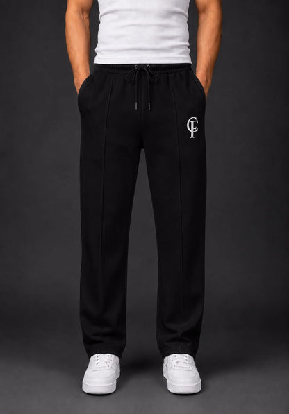Black Trouser with a white DTF FlexaClub "FC" monogram logo on the left thigh, modeled with a white tank top and white sneakers against a dark gray studio background.