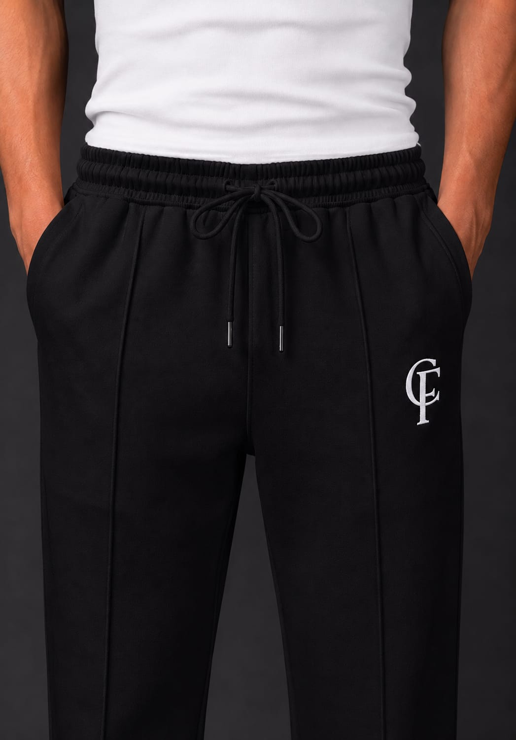 Black Trouser with a white DTF FlexaClub "FC" monogram logo on the left thigh, modeled with a white tank top and white sneakers against a dark gray studio background.