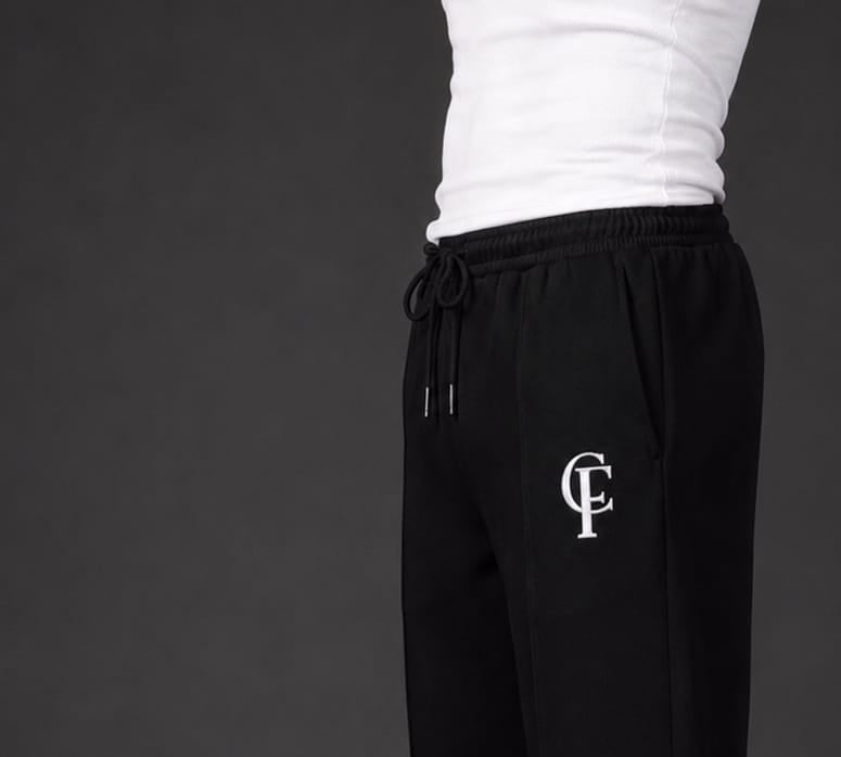 Black Trouser with a white DTF FlexaClub "FC" monogram logo on the left thigh, modeled with a white tank top and white sneakers against a dark gray studio background.