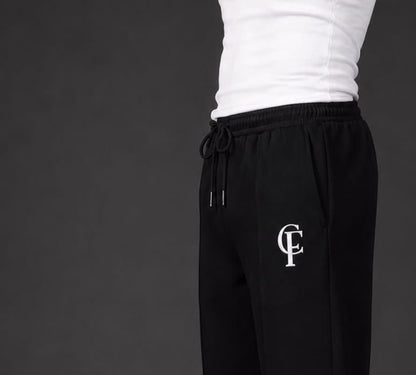 Black Trouser with a white DTF FlexaClub "FC" monogram logo on the left thigh, modeled with a white tank top and white sneakers against a dark gray studio background.