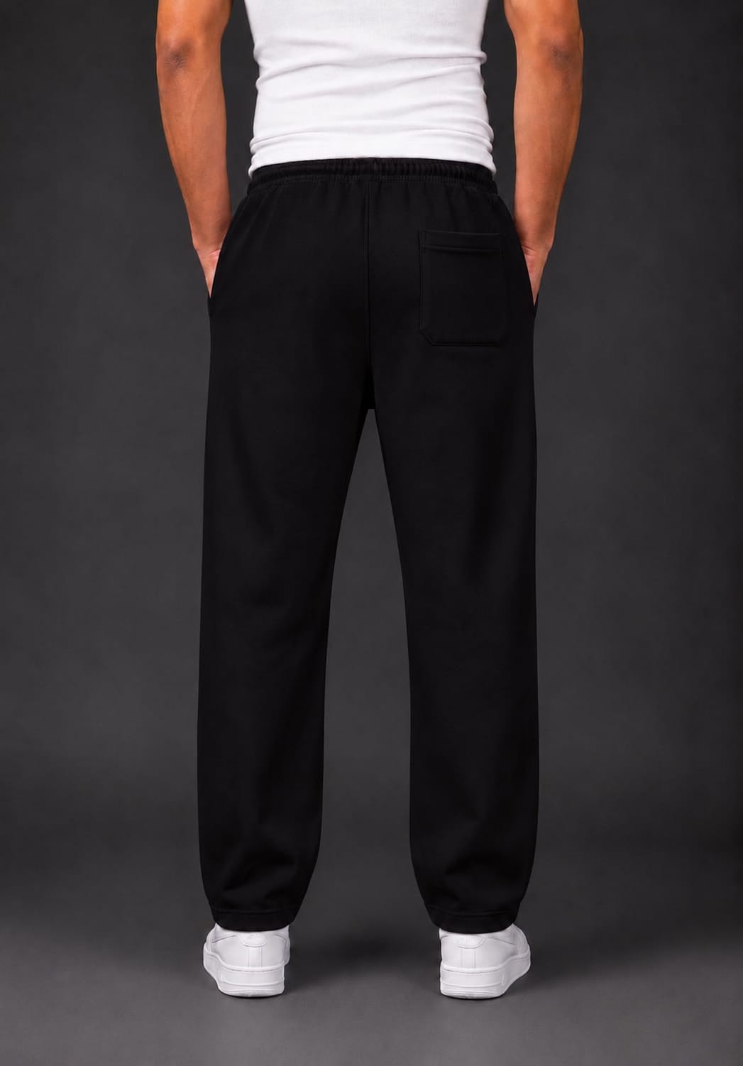 A person shown from the back wearing loose black trouser with a side back pocket, a white tank top, and white sneakers against a dark gray studio background.