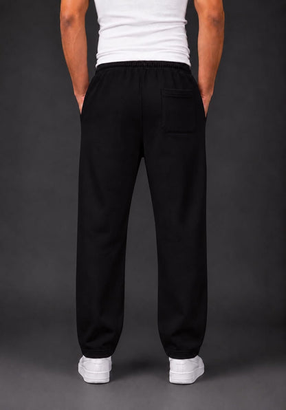 A person shown from the back wearing loose black trouser with a side back pocket, a white tank top, and white sneakers against a dark gray studio background.