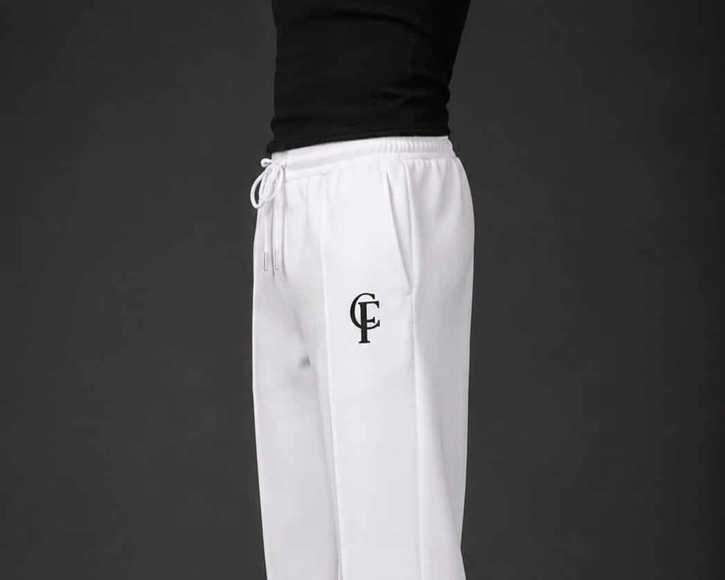 White Trouser with a black DTF FlexaClub "FC" monogram logo on the left thigh, modeled with a black tank top and white sneakers against a dark gray studio background.