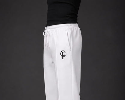 White Trouser with a black DTF FlexaClub "FC" monogram logo on the left thigh, modeled with a black tank top and white sneakers against a dark gray studio background.