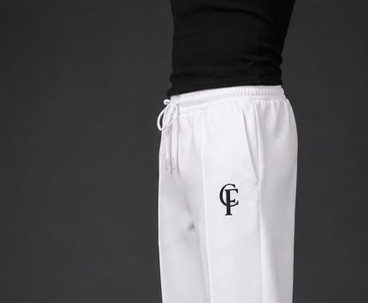 White sweatpants with a black embroidered monogram logo on the left thigh, worn by a model with a black top and white sneakers against a dark gray studio background.