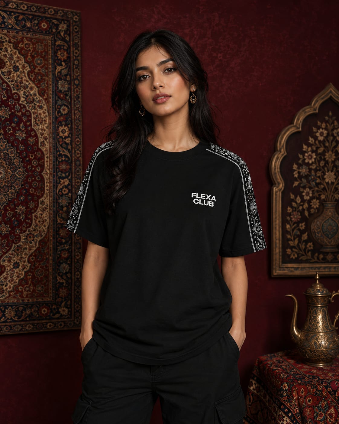 A woman modeling a black drop shoulder T-shirt with white 'FLEXA CLUB' text on the chest and patterned sleeve details, set against a dark red wall with traditional patterns.