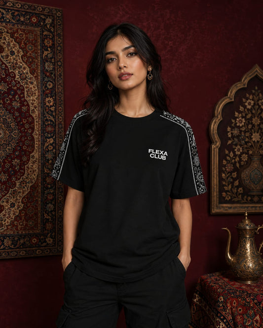 A woman modeling a black drop shoulder T-shirt with white 'FLEXA CLUB' text on the chest and patterned sleeve details, set against a dark red wall with traditional patterns.