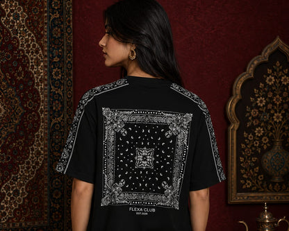 A woman shown from the back wearing a black drop shoulder T-shirt with a large white bandana-style graphic print on the back and white patterned shoulder straps. She is standing in a dark red interior setting with ornate rugs and wall decor.