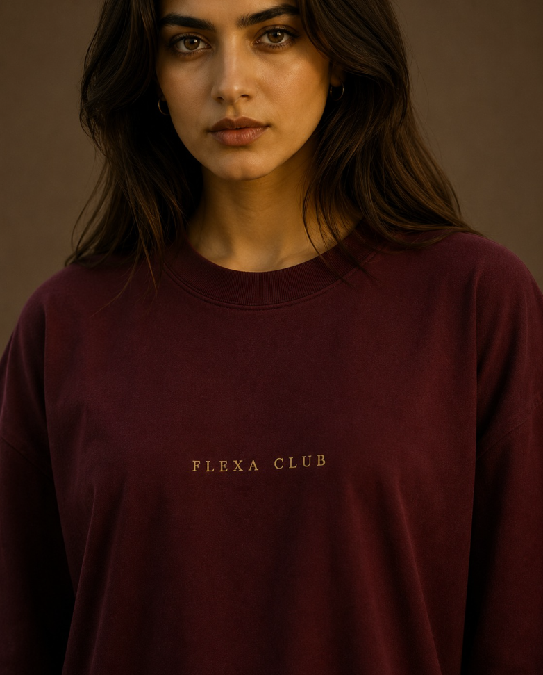 A woman wearing a dark burgundy crewneck drop shoulder T-Shirt with 'FLEXA CLUB' printed in gold lettering on the chest, set against a warm brown background.