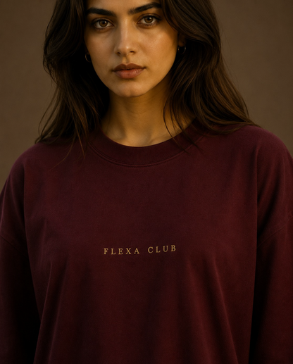 A woman wearing a dark burgundy crewneck drop shoulder T-Shirt with 'FLEXA CLUB' printed in gold lettering on the chest, set against a warm brown background.