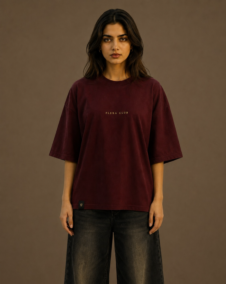 A woman modeling an oversized maroon drop shoulder T-shirt with 'FLEKA CLUB' printed in small gold letters on the chest, paired with dark gray jeans, against a solid light brown studio background.