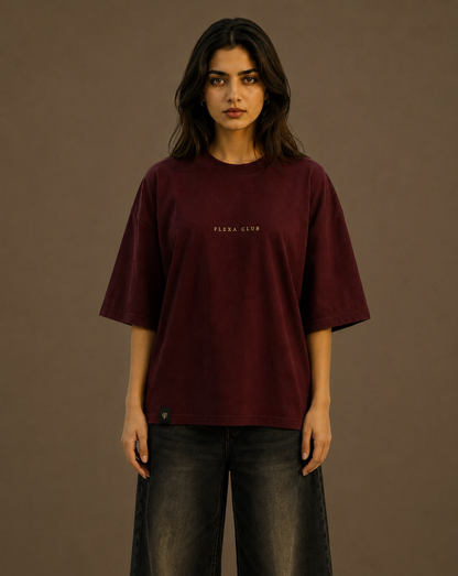 A woman modeling an oversized maroon drop shoulder T-shirt with 'FLEKA CLUB' printed in small gold letters on the chest, paired with dark gray jeans, against a solid light brown studio background.