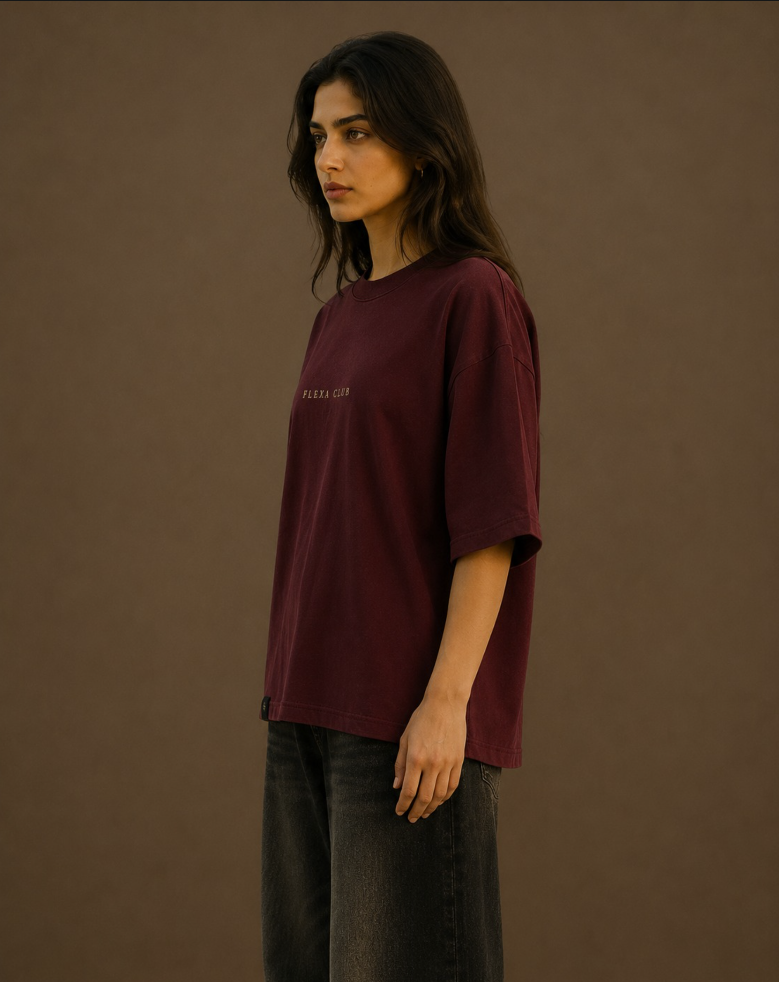 A woman wearing an oversized maroon drop shoulder T-shirt with small white text on the chest, paired with dark gray jeans, standing against a solid brown studio background.
