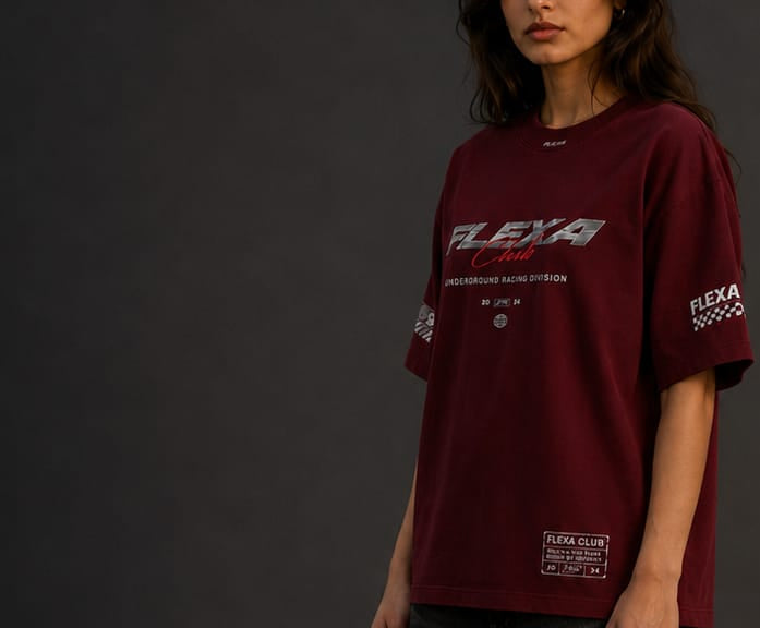 A woman wearing an oversized maroon t-shirt with 'FLEXA Club' racing graphics, shown from the chest down against a dark gray studio background.
