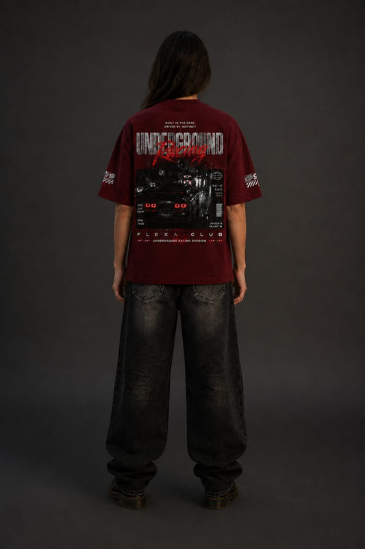 A woman wearing an oversized maroon t-shirt with 'FLEXA Club' with faded black baggy jeans in street racing graphics, shown from the back against a dark gray studio background.