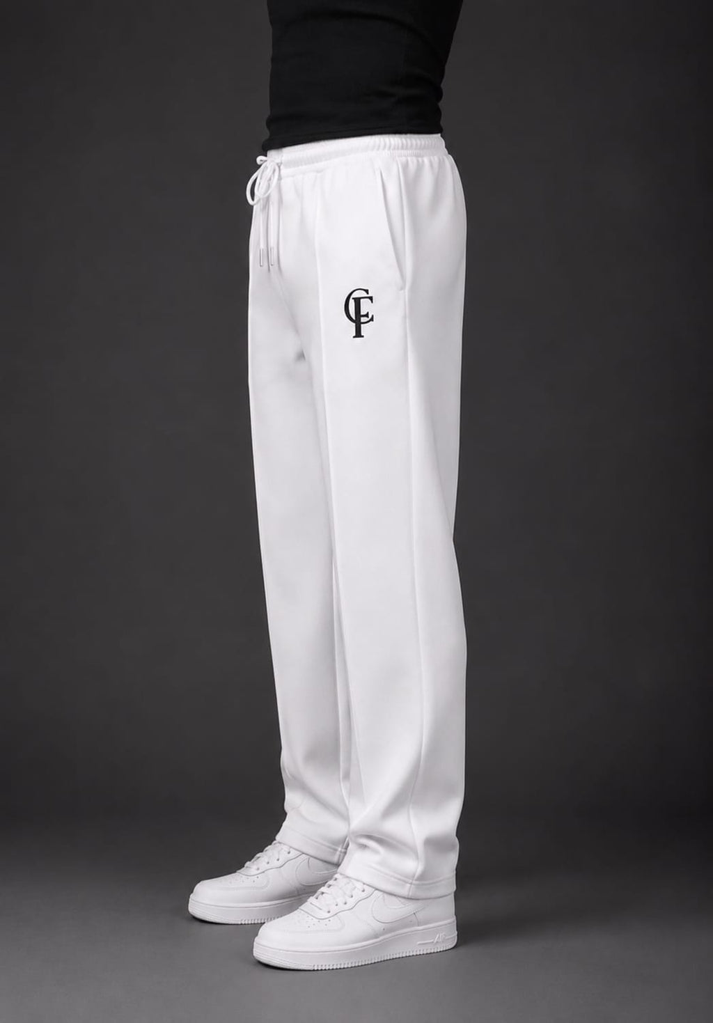 White sweatpants with a black embroidered monogram logo on the left thigh, worn by a model with a black top and white sneakers against a dark gray studio background.
