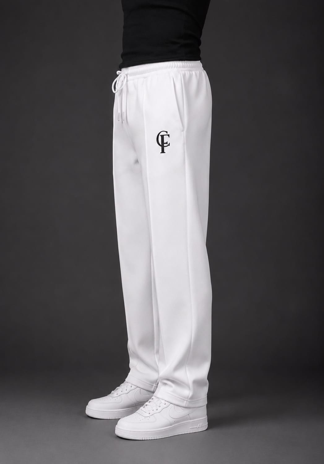 White sweatpants with a black embroidered monogram logo on the left thigh, worn by a model with a black top and white sneakers against a dark gray studio background.
